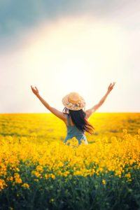 Back Silhouette Woman raised her hands to sky, sun, sunset. Blue dress straw trendy hat. Field rape yellow flowers. Long hair brunette fly in motion wind. Concept religion prayer blessing divine light